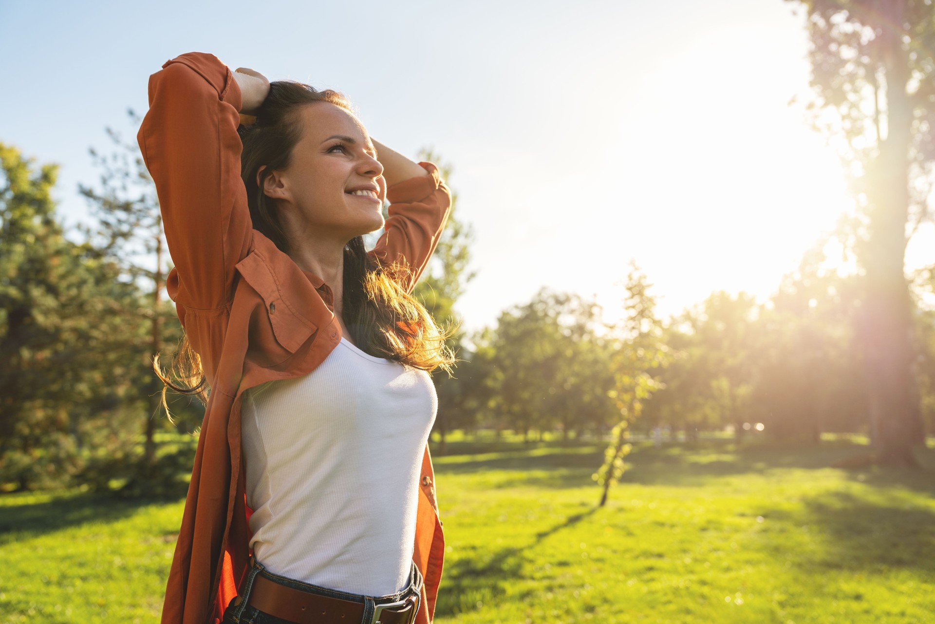 Optimistic Caucasian young woman enjoying springtime outdoors walking in city park. Woman stands with hands behind head and smiling.