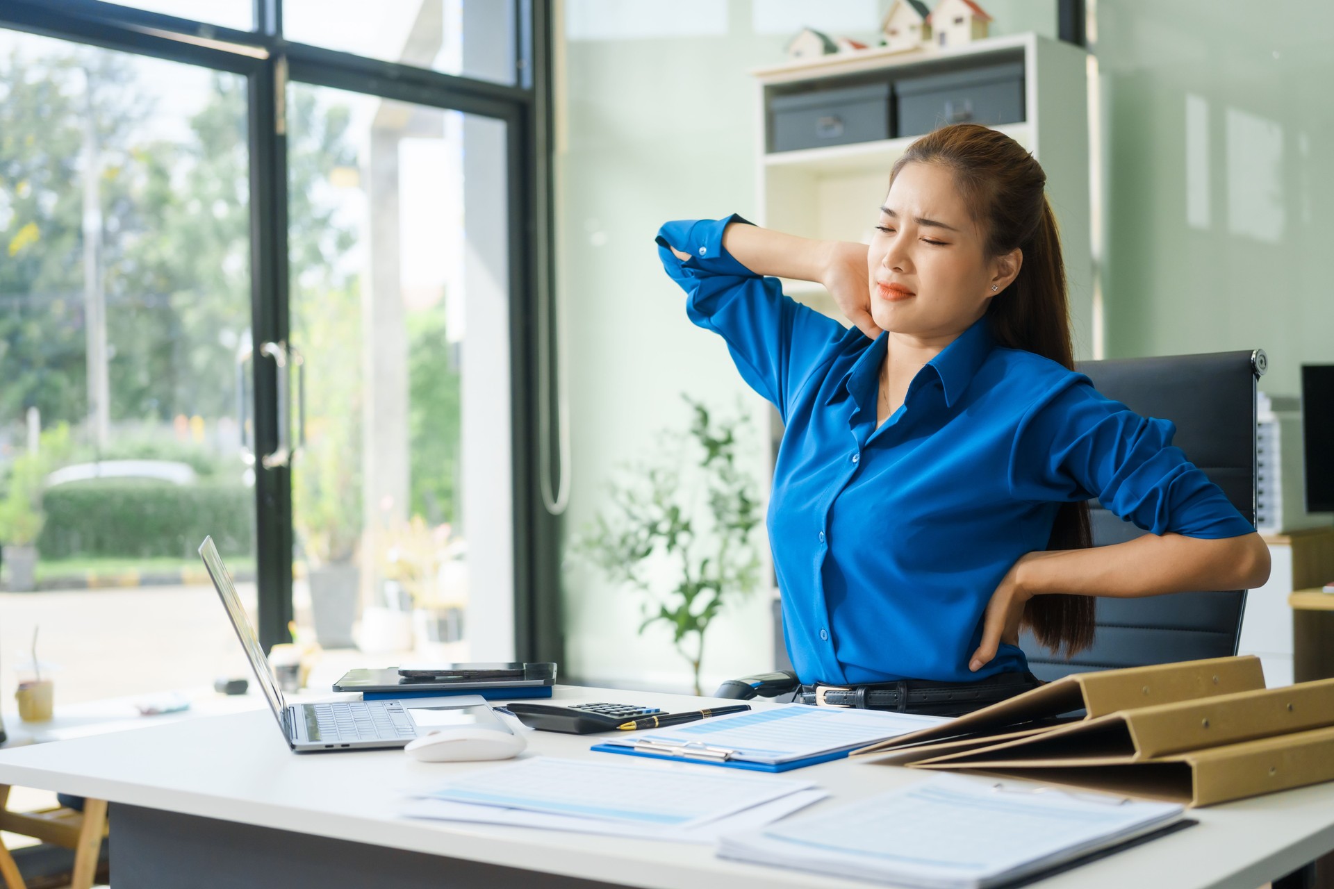 A businesswoman working at a desk in an office struggles with stress and headaches caused by Office Syndrome, a condition linked to prolonged sitting excessive computer use during long working hours