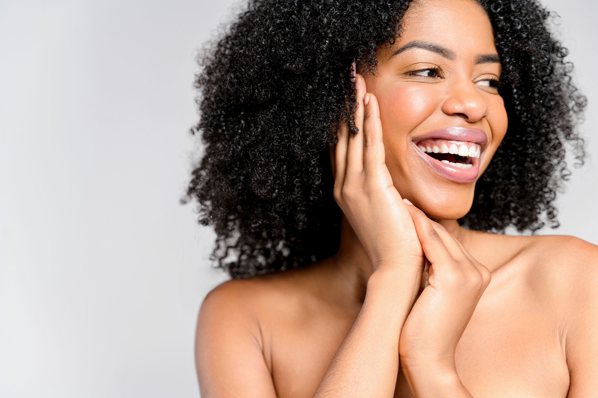 A joyful African-American woman with a vivacious smile and natural curly hair poses with her hands gently framing her face